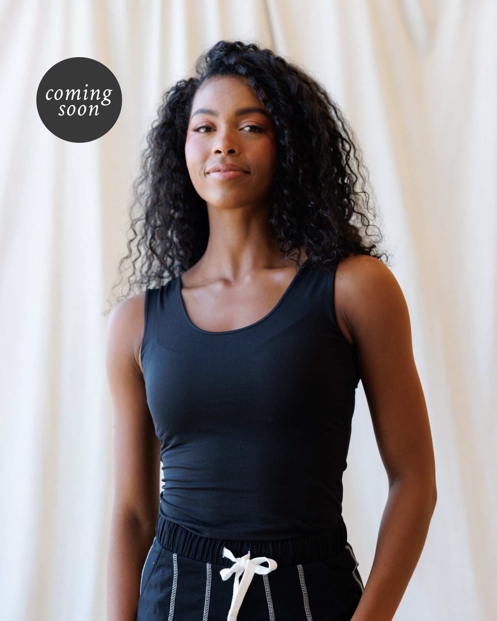 A woman with curly dark hair wearing a black sleeveless scoop-neck top and black pants with a white drawstring, standing against a draped cream fabric backdrop. A circular 'coming soon' badge appears in the upper left.