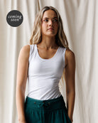 A woman wearing a fitted white scoop-neck tank top and dark teal drawstring pants, standing against a draped cream fabric backdrop. A black circular 'coming soon' badge appears in the upper left.