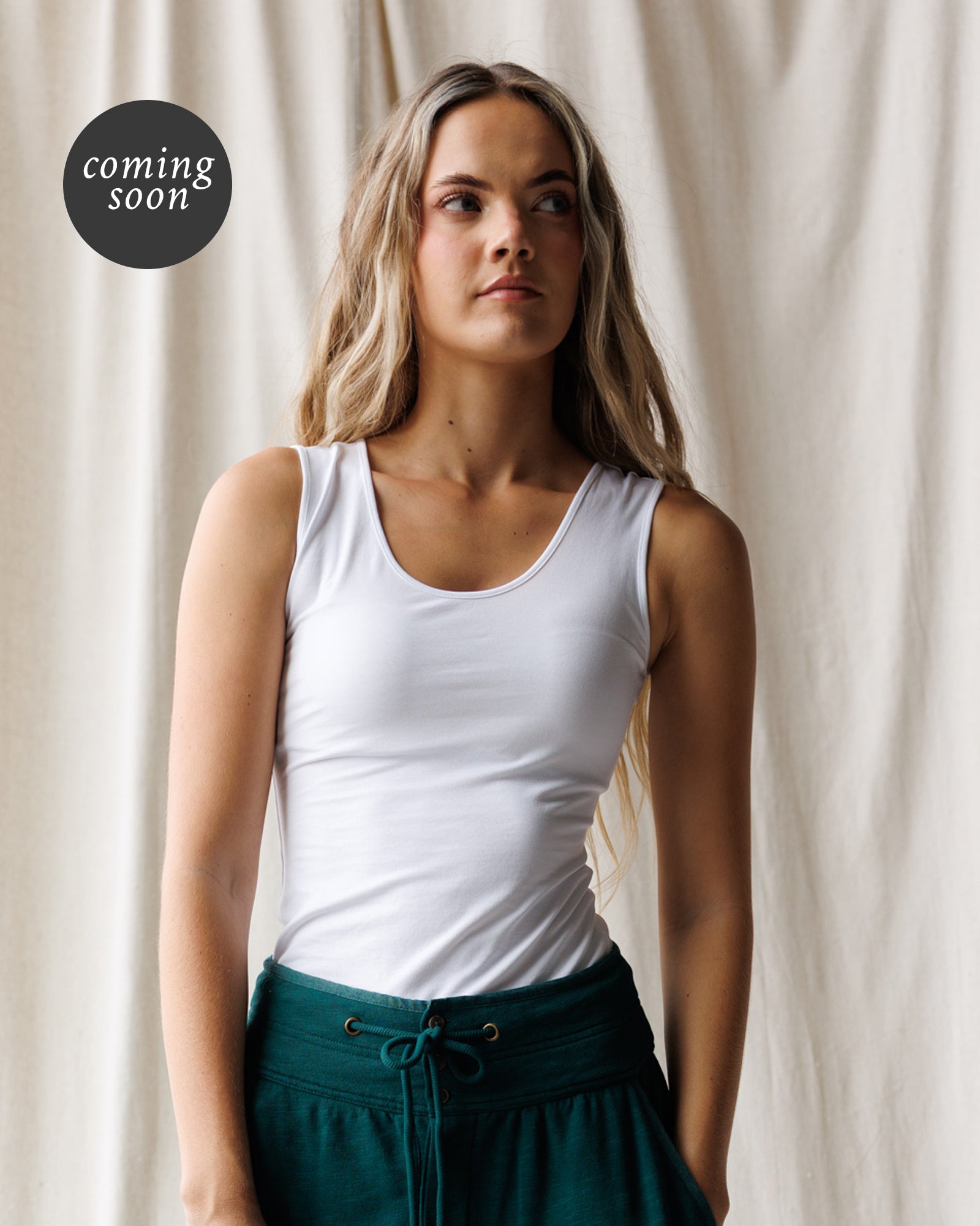 A woman wearing a fitted white scoop-neck tank top and dark teal drawstring pants, standing against a draped cream fabric backdrop. A black circular 'coming soon' badge appears in the upper left.