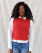 A woman with long, curly black hair wearing a red cable-knit sweater vest over a white collared shirt and blue jeans, standing against a draped white fabric backdrop.