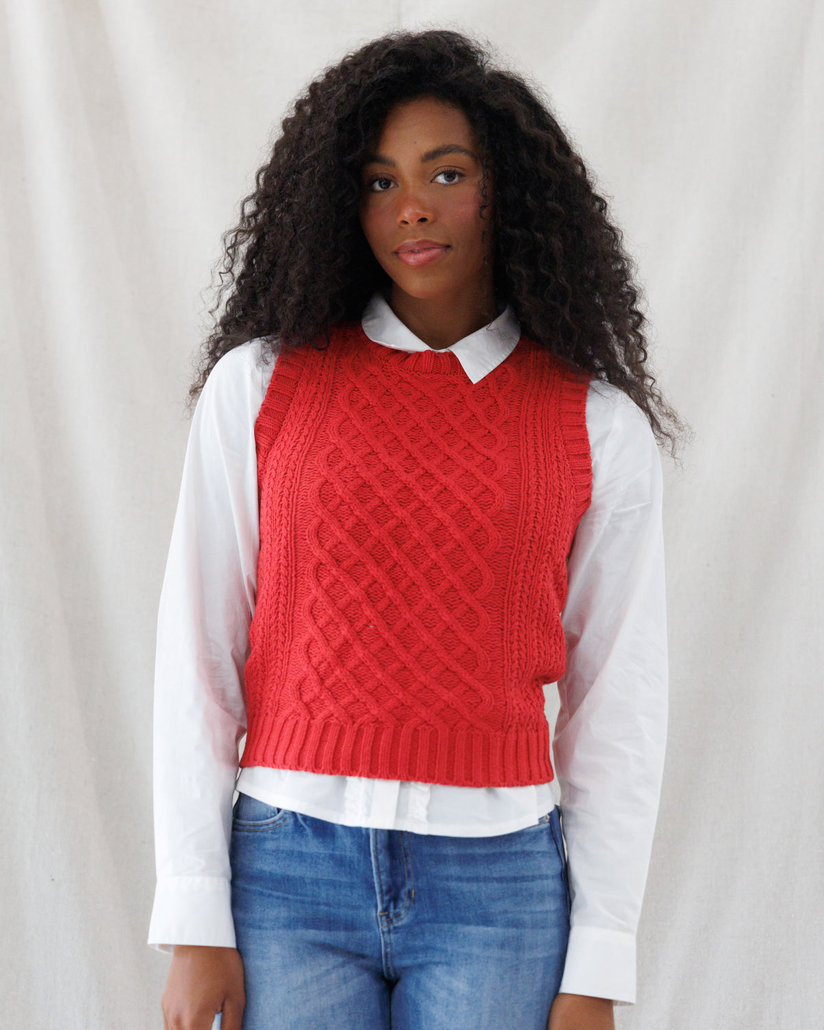 A woman with long, curly black hair wearing a red cable-knit sweater vest over a white collared shirt and blue jeans, standing against a draped white fabric backdrop.
