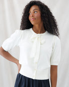 A woman with curly dark hair wearing a cream short-sleeve knit top with a large bow at the collar and a dark skirt, posed against a light draped fabric backdrop.