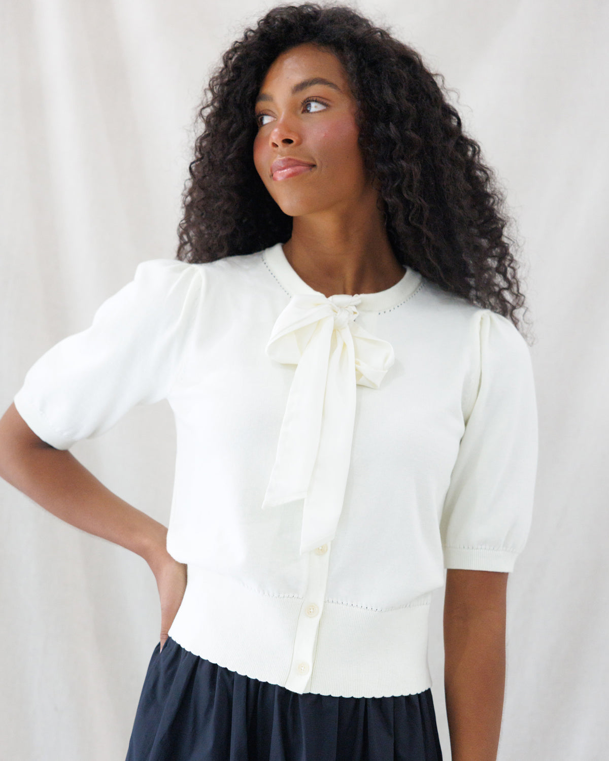 A woman with curly dark hair wearing a cream short-sleeve knit top with a large bow at the collar and a dark skirt, posed against a light draped fabric backdrop.