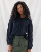 A woman with long, curly dark hair wearing a black long-sleeve blouse with vertical pleating and a high neckline, paired with an olive green skirt, standing against a white draped fabric background.