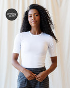 A woman with long, curly black hair is wearing a fitted white short-sleeve crewneck top and dark patterned pants, standing against a draped cream fabric backdrop. A black circular 'coming soon' badge is in the upper left corner.