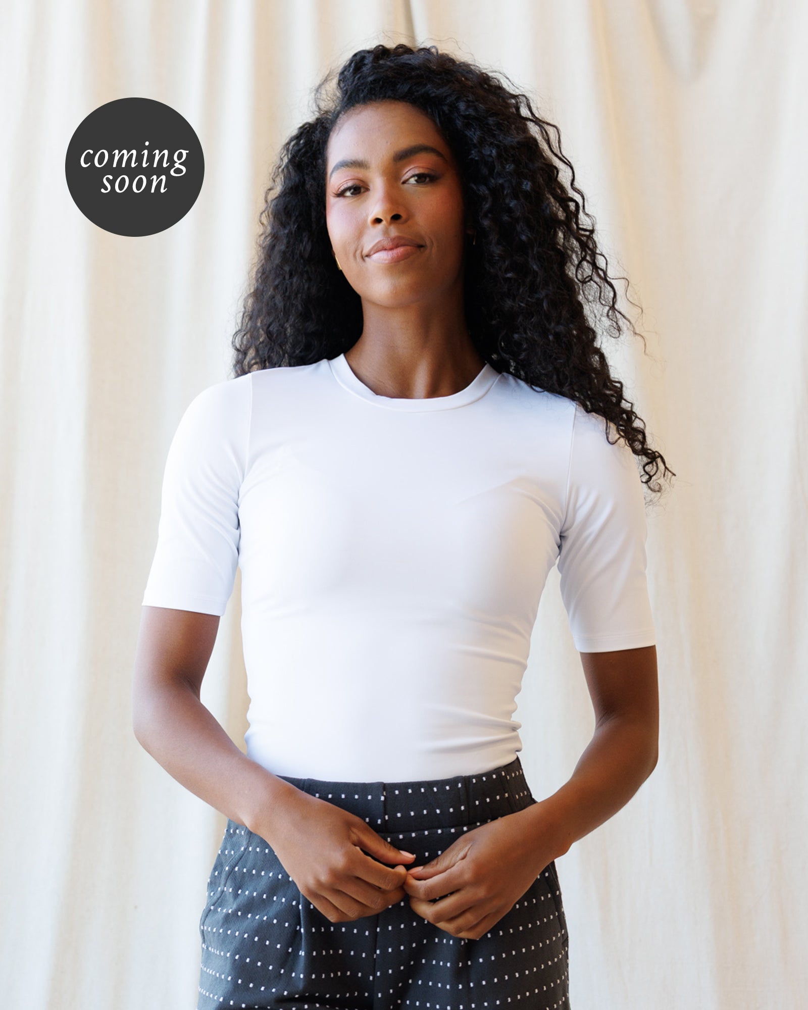 A woman with long, curly black hair is wearing a fitted white short-sleeve crewneck top and dark patterned pants, standing against a draped cream fabric backdrop. A black circular 'coming soon' badge is in the upper left corner.