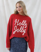 A woman wearing a red knit sweater with 'Holly Jolly' written in white script across the front, paired with a sparkly navy skirt, posed against a draped white fabric backdrop.