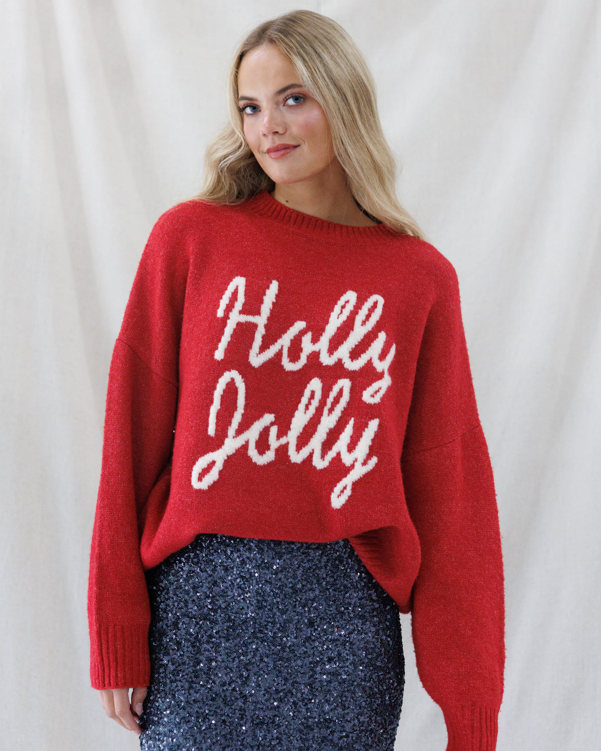 A woman wearing a red knit sweater with 'Holly Jolly' written in white script across the front, paired with a sparkly navy skirt, posed against a draped white fabric backdrop.