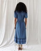 A woman viewed from behind wearing a blue denim midi dress with short puff sleeves, a gathered waist, and a tiered ruffle hem, standing against a white fabric backdrop.