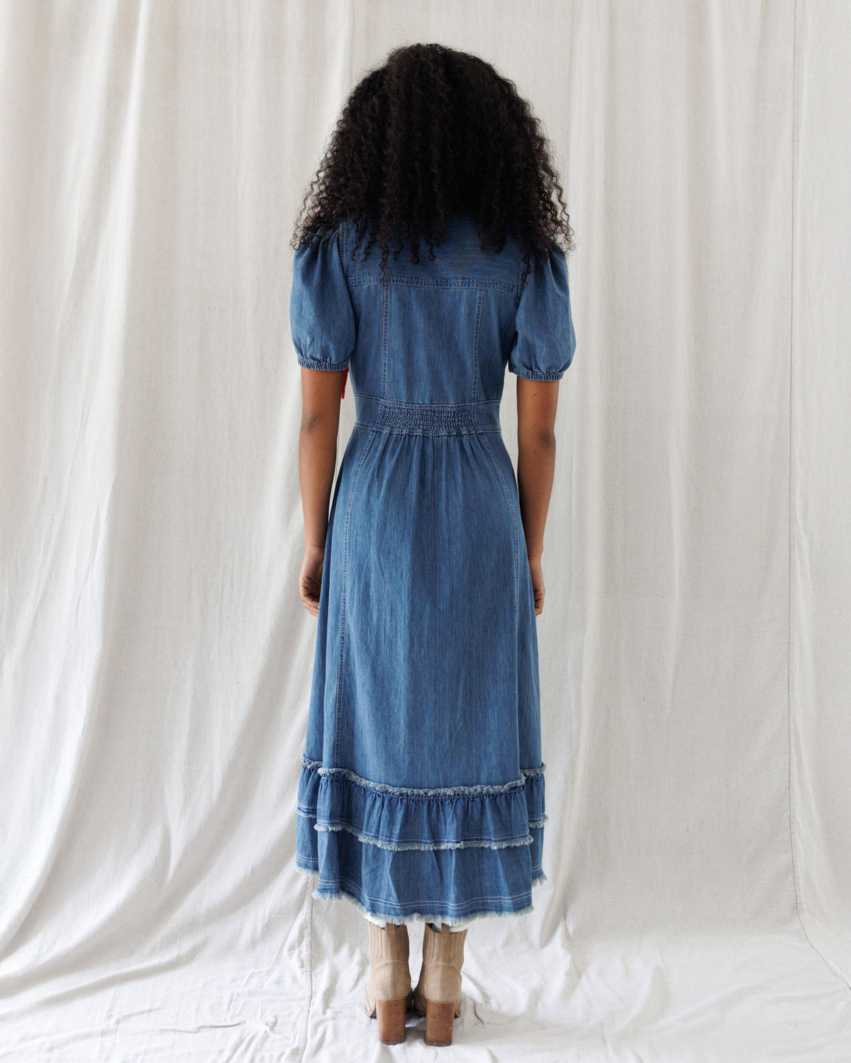 A woman viewed from behind wearing a blue denim midi dress with short puff sleeves, a gathered waist, and a tiered ruffle hem, standing against a white fabric backdrop.