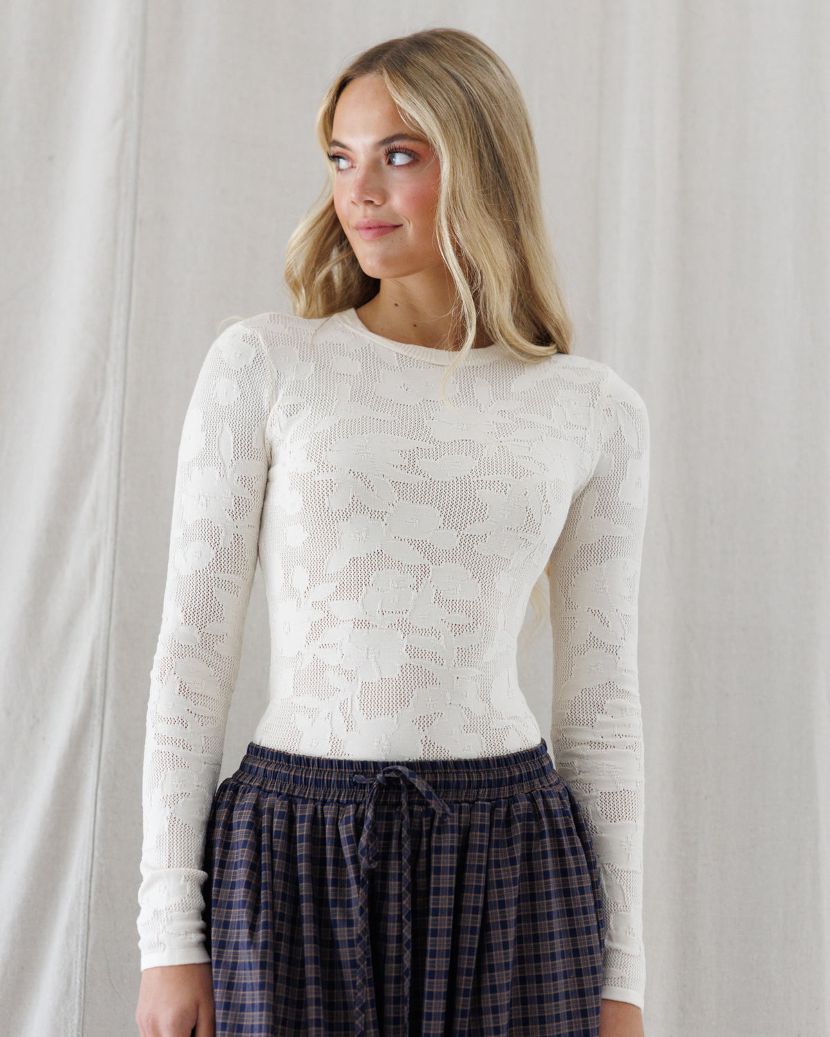 A woman wearing a fitted, long-sleeve cream lace top and dark plaid pants, standing against a draped white fabric backdrop.