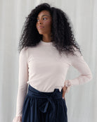 A woman with long, curly black hair wearing a light pink ribbed long-sleeve mock neck top and a navy blue skirt with a tie waist, standing against a light gray curtain background.