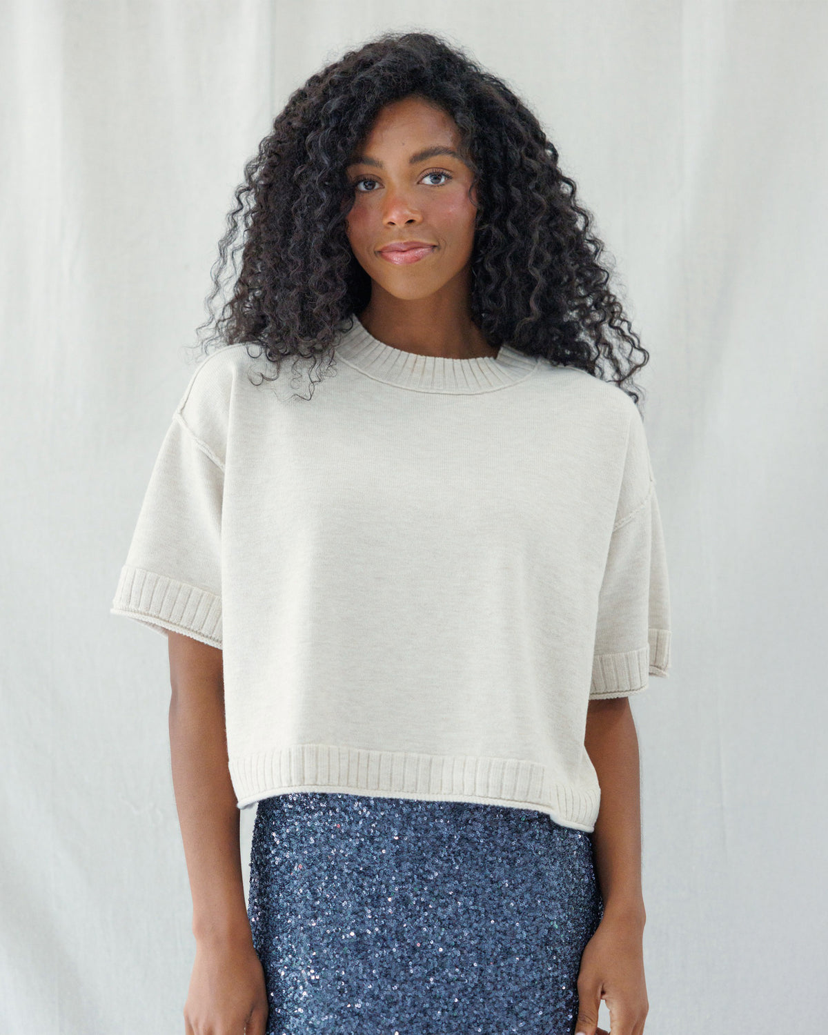 A woman with curly black hair wearing a cream-colored short-sleeve knit sweater and a blue sequined skirt, standing against a white fabric backdrop.