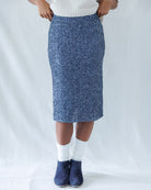 A person wearing a blue speckled knit pencil skirt with a white ribbed top, white socks, and navy blue slip-on shoes, standing against a white fabric backdrop.