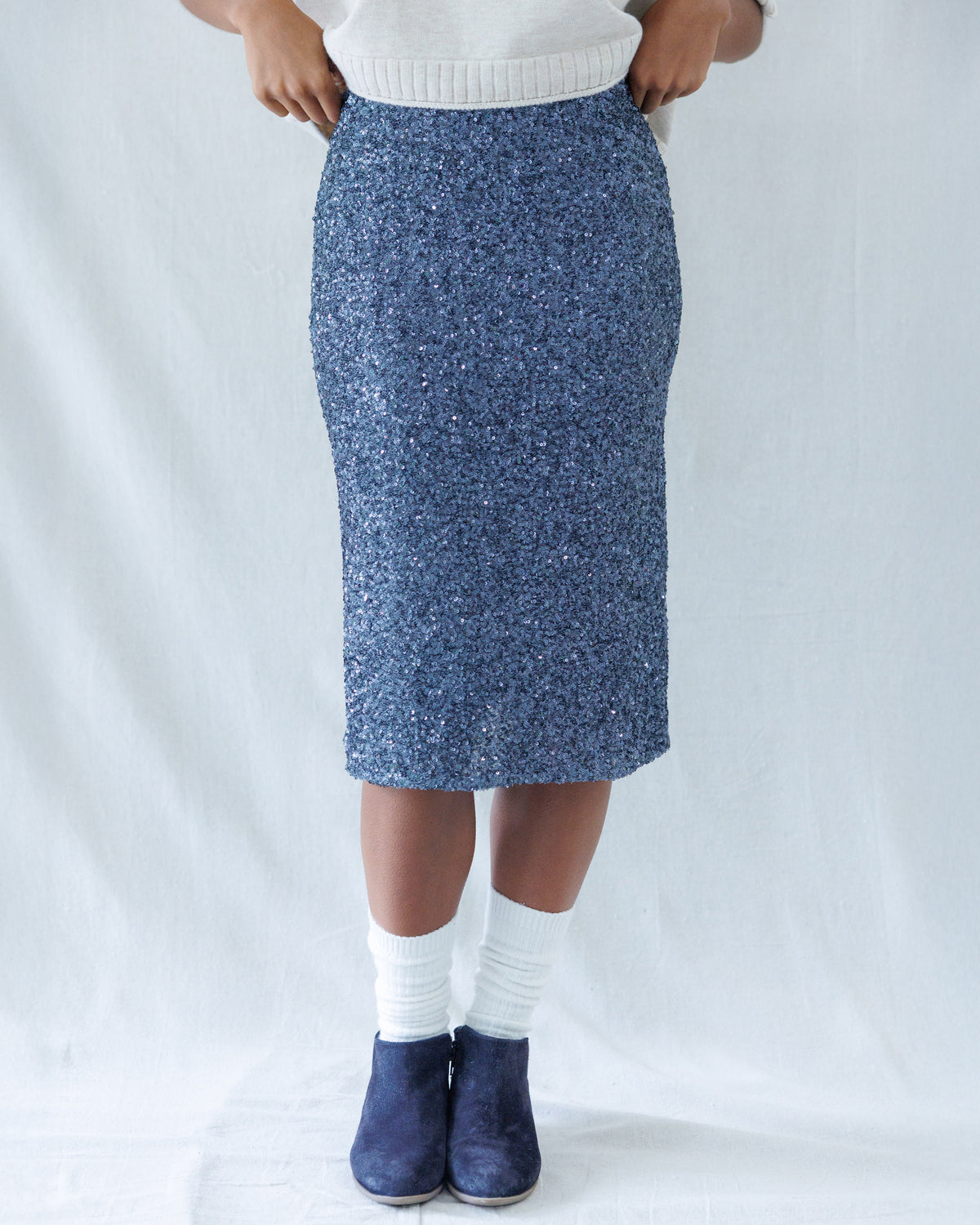 A person wearing a blue speckled knit pencil skirt with a white ribbed top, white socks, and navy blue slip-on shoes, standing against a white fabric backdrop.