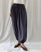 A person wearing loose, high-waisted harem-style pants with a dark blue and black checkered pattern, paired with a white long-sleeve top and brown slip-on shoes, standing against a white fabric backdrop.