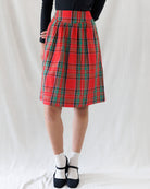 A red and green tartan plaid pleated skirt worn by a model, paired with a black long-sleeve top, white socks, and black Mary Jane shoes, photographed against a white fabric backdrop.