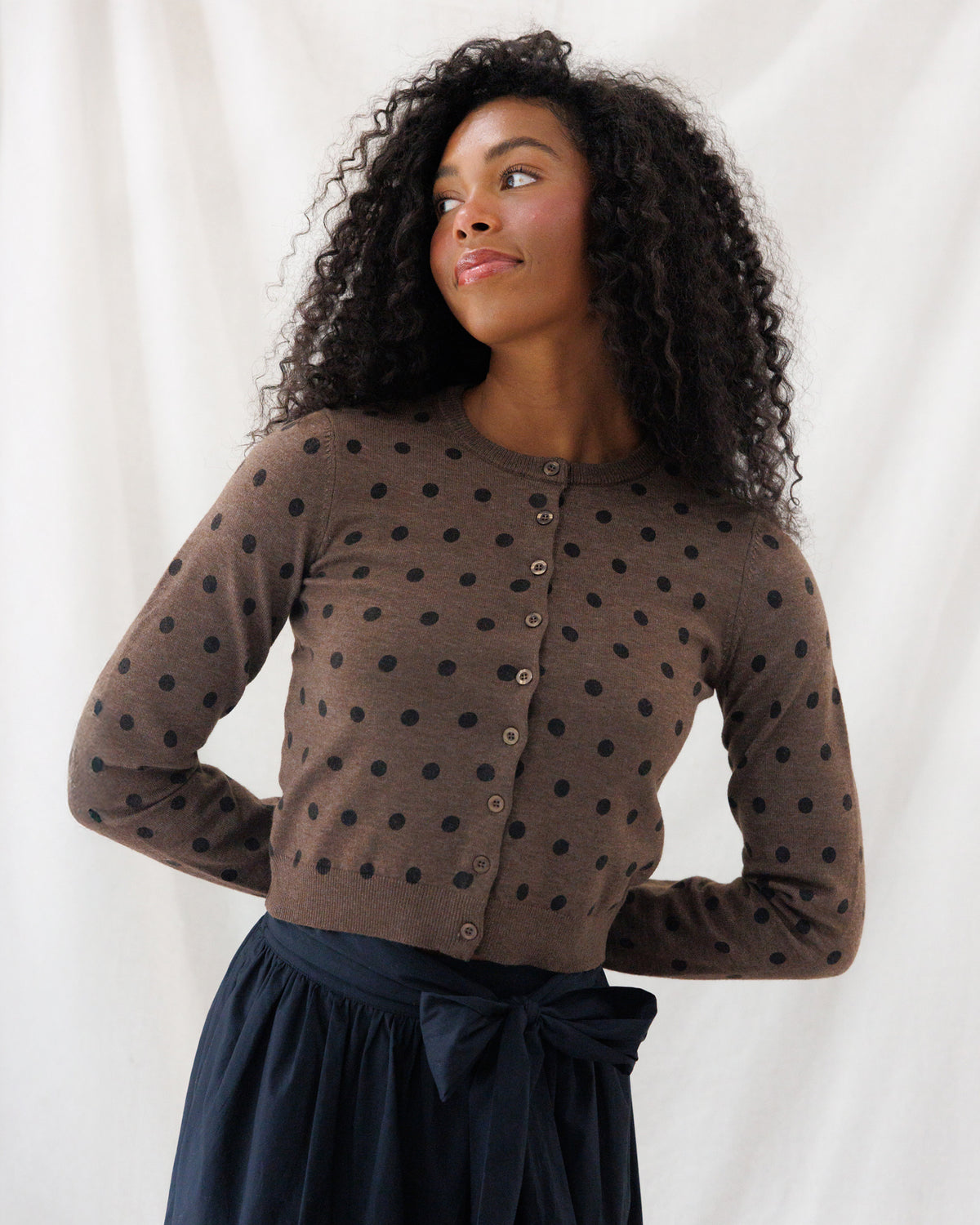 A woman with curly black hair wearing a brown button-front cardigan with black polka dots and a navy skirt, posing against a white draped fabric background.