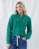 A woman wearing a vibrant green cable-knit cardigan sweater with brown buttons, paired with blue jeans, standing against a white fabric backdrop.