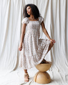 Woman in a floral dress standing on a wooden stool against a white curtain background