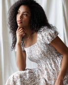 Woman wearing a white and brown floral dress sitting against a white curtain