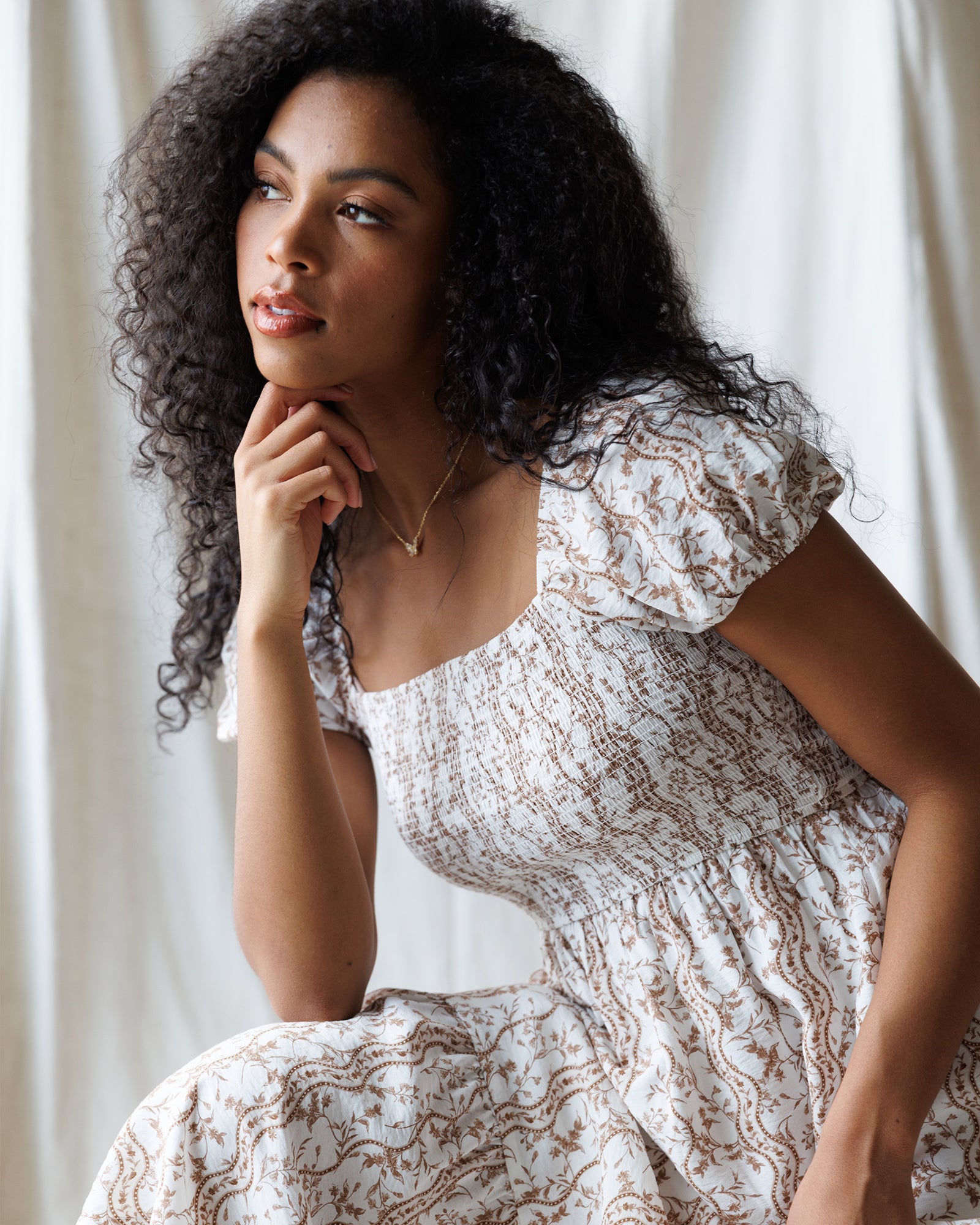 Woman wearing a white and brown floral dress sitting against a white curtain
