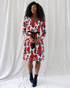 A woman modeling a white knee-length dress with a vibrant red, pink, and green floral print, featuring a black waist tie belt, standing against a white draped fabric backdrop.