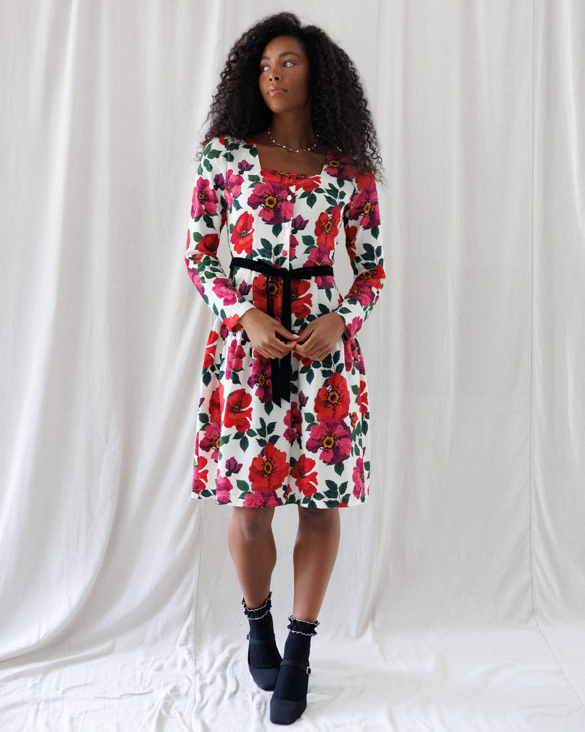 A woman modeling a white knee-length dress with a vibrant red, pink, and green floral print, featuring a black waist tie belt, standing against a white draped fabric backdrop.