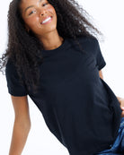 A smiling woman with long curly black hair wearing a plain black short-sleeve crewneck t-shirt and blue jeans, photographed against a clean white background.